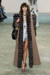 Runway model wearing a long quilted beige cape with thick fur trim, white slip dress, and purple heels, carrying a handbag.