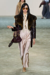 Model walking a runway in a cream satin jumpsuit with lace blouse, a dark fur stole, and a burgundy leather coat; she wears brown aviator sunglasses and carries a burgundy handbag.