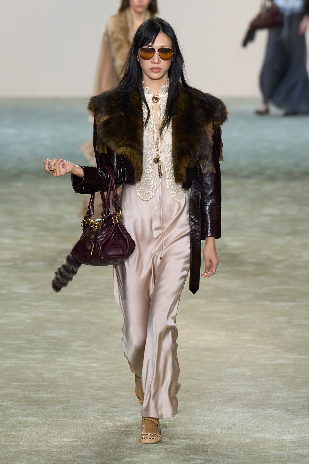 Model walking a runway in a cream satin jumpsuit with lace blouse, a dark fur stole, and a burgundy leather coat; she wears brown aviator sunglasses and carries a burgundy handbag.