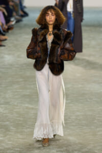 Female model walking a runway in an oversized brown fur-trimmed leather coat over a long white lace dress with a slit at the side.