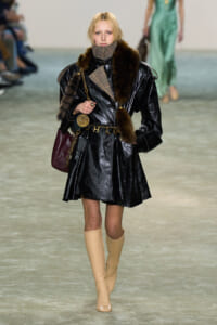 Model walks the runway in a shiny black patent trench with fur trim, plaid scarf, beige knee-high boots, and a burgundy handbag.