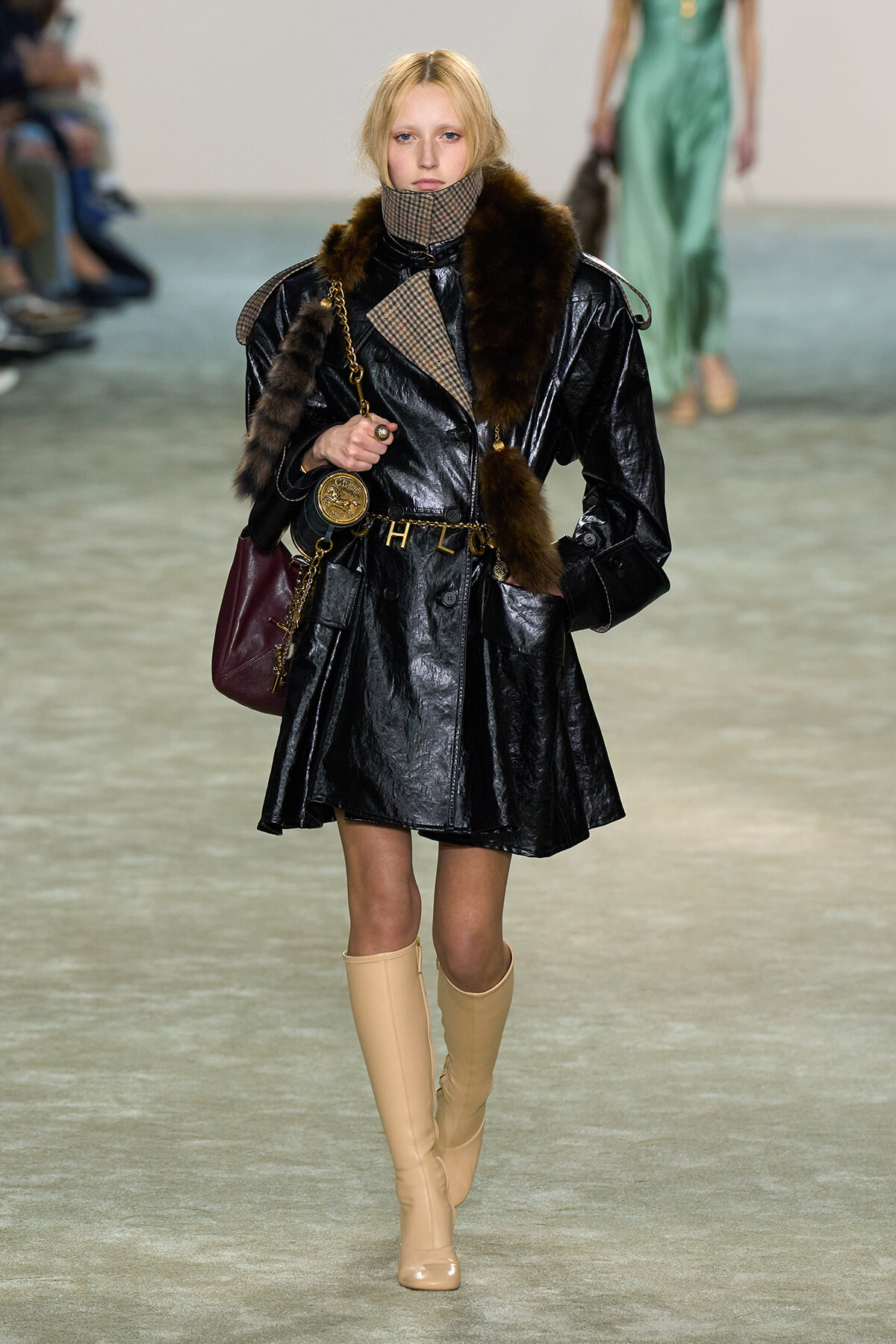 Model walks the runway in a shiny black patent trench with fur trim, plaid scarf, beige knee-high boots, and a burgundy handbag.