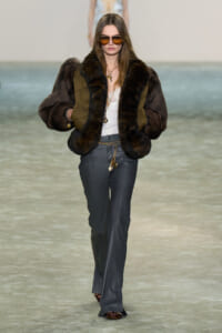 Model walking runway in a brown fur-trimmed jacket with olive panels, gray leather pants, and brown sunglasses