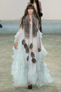 Model walking a runway in a pale mint-green, tiered chiffon gown with a gray fur stole and gold chain accents with fur pom-poms hanging from the chains.