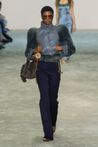 Model on a runway wearing a blue-gray faux-fur bolero over a metallic gray high-collar jacket, navy pants, and brown shoes with gold accessories.