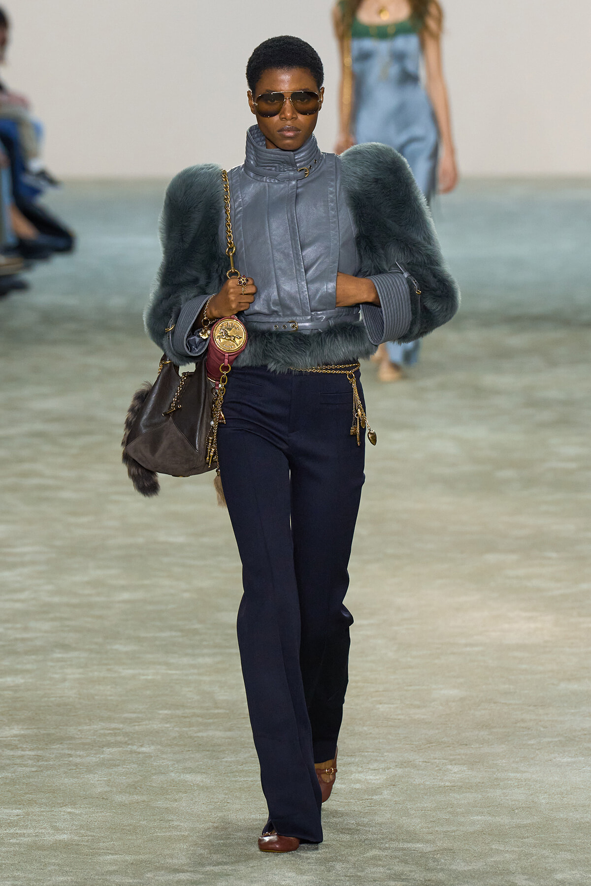 Model on a runway wearing a blue-gray faux-fur bolero over a metallic gray high-collar jacket, navy pants, and brown shoes with gold accessories.