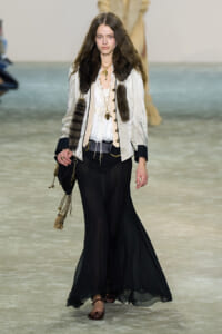 Model walking the runway in a cream jacket with fur trim over a lace blouse, black wide-leg sheer pants, and layered gold jewelry; brown flats and a chain-adorned bag