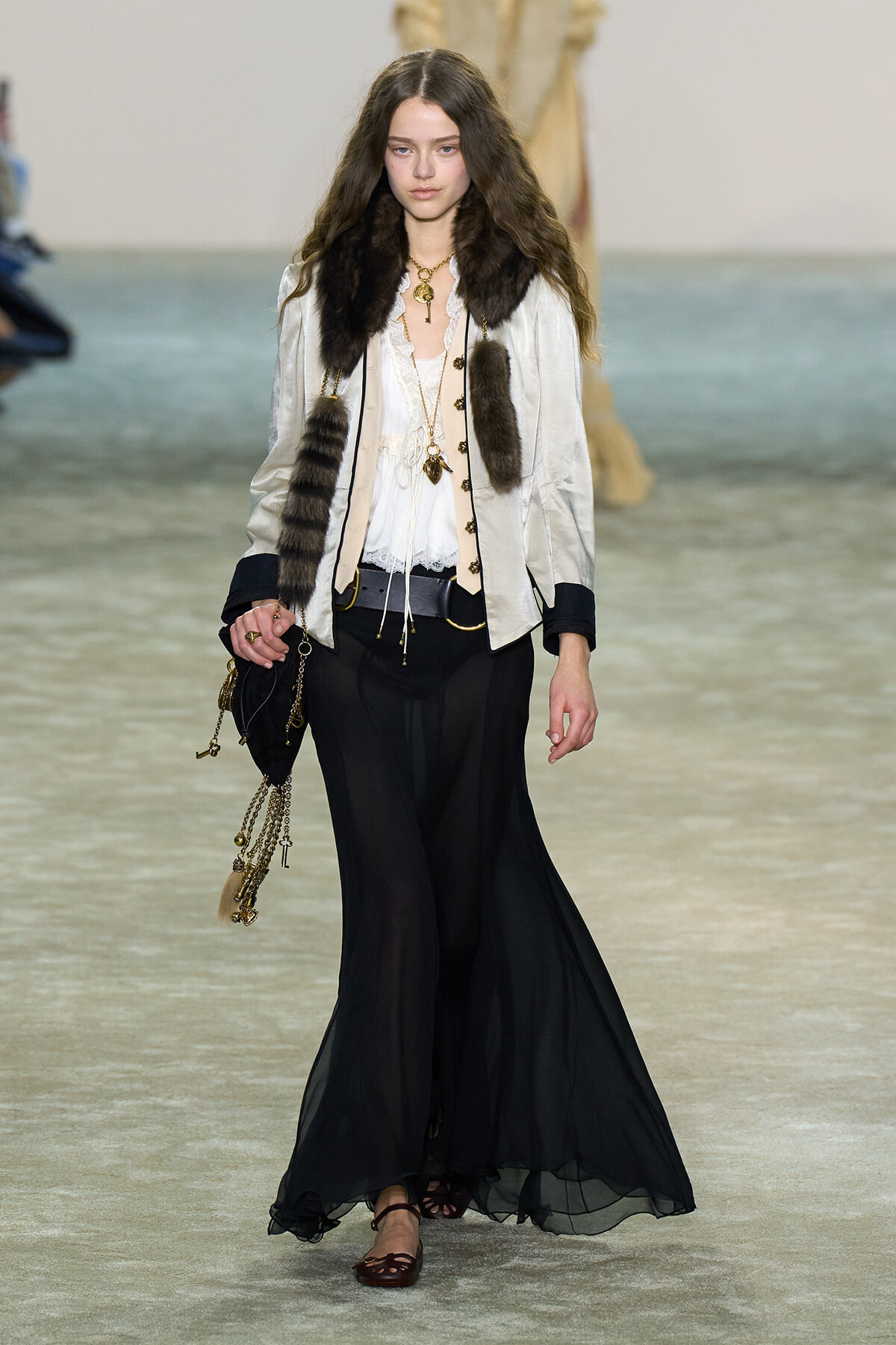 Model walking the runway in a cream jacket with fur trim over a lace blouse, black wide-leg sheer pants, and layered gold jewelry; brown flats and a chain-adorned bag
