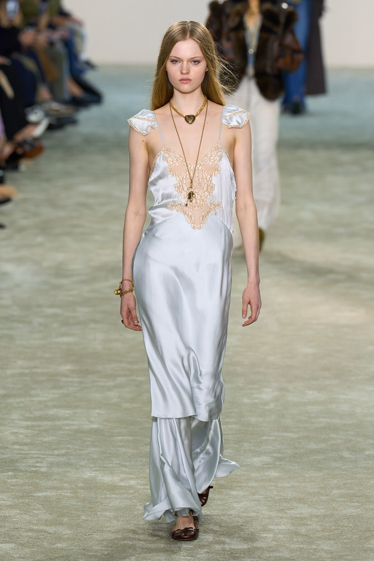 Model walks the runway in a pale blue satin slip dress with peach lace bodice and layered skirt, accessorized with gold jewelry.
