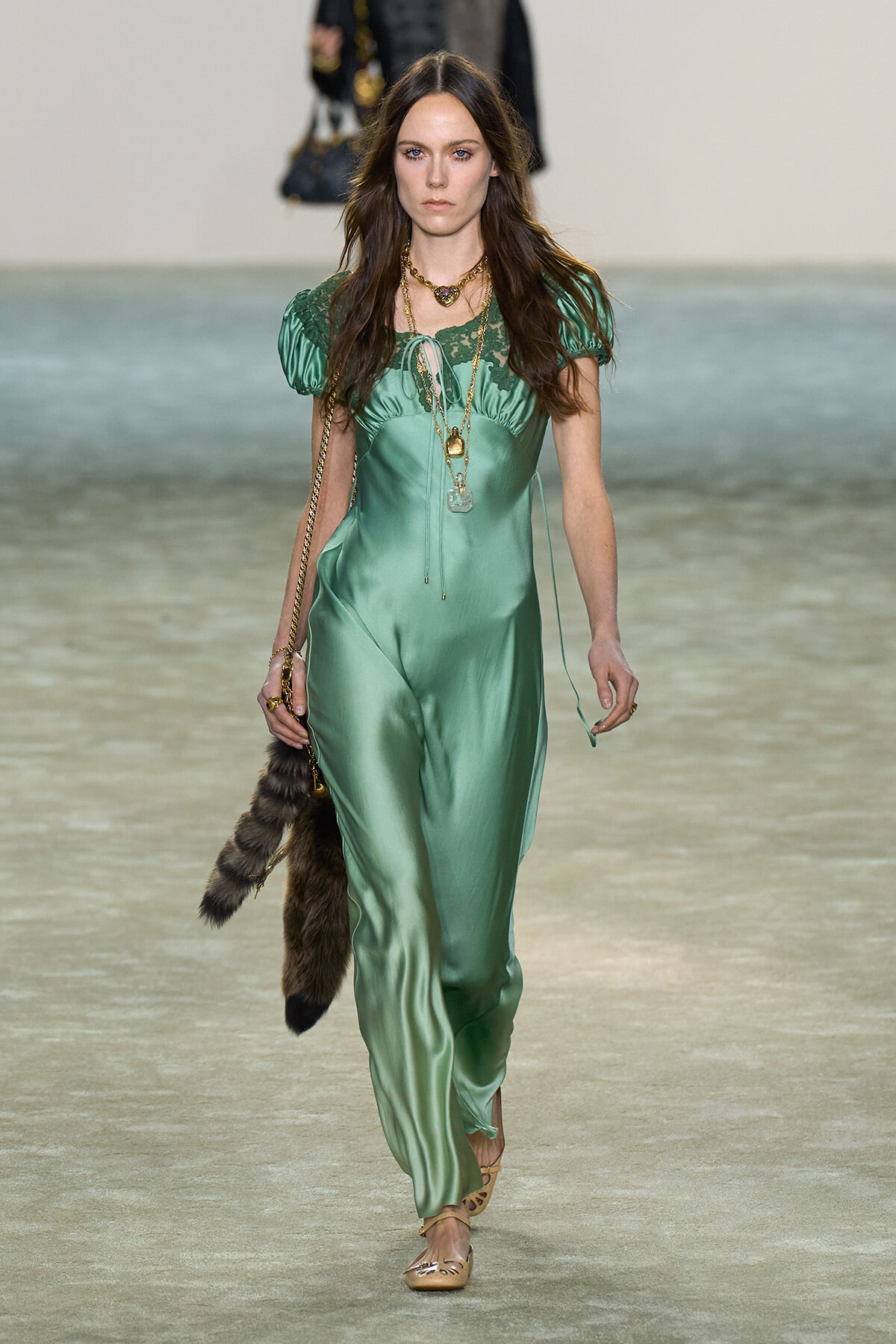 Runway model in emerald-green satin jumpsuit with lace bodice and puff sleeves, layered gold necklaces, and a fur accessory hanging from her hand.
