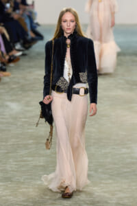 Model walks the runway in a dark velvet jacket with jeweled pockets, paired with a flowing cream skirt and gold accessories.