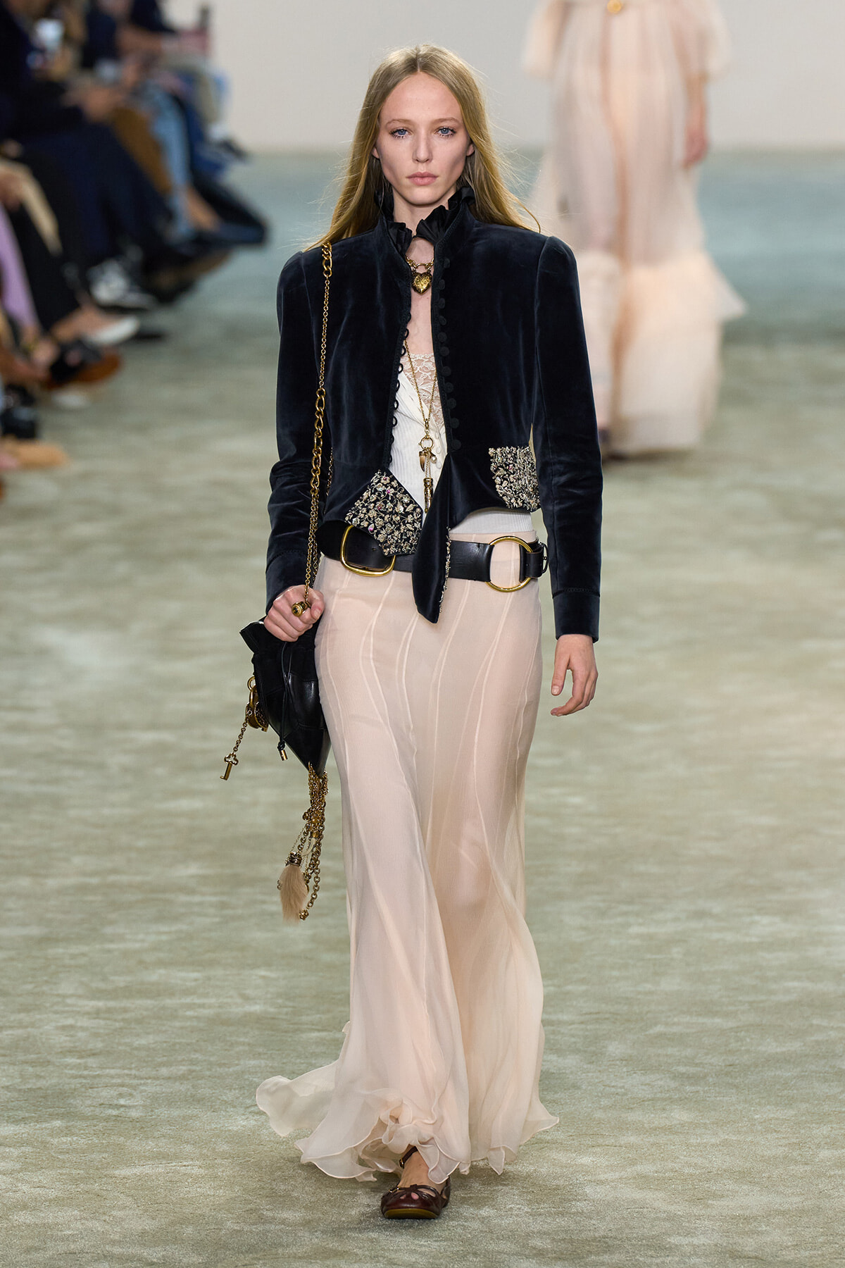 Model walks the runway in a dark velvet jacket with jeweled pockets, paired with a flowing cream skirt and gold accessories.