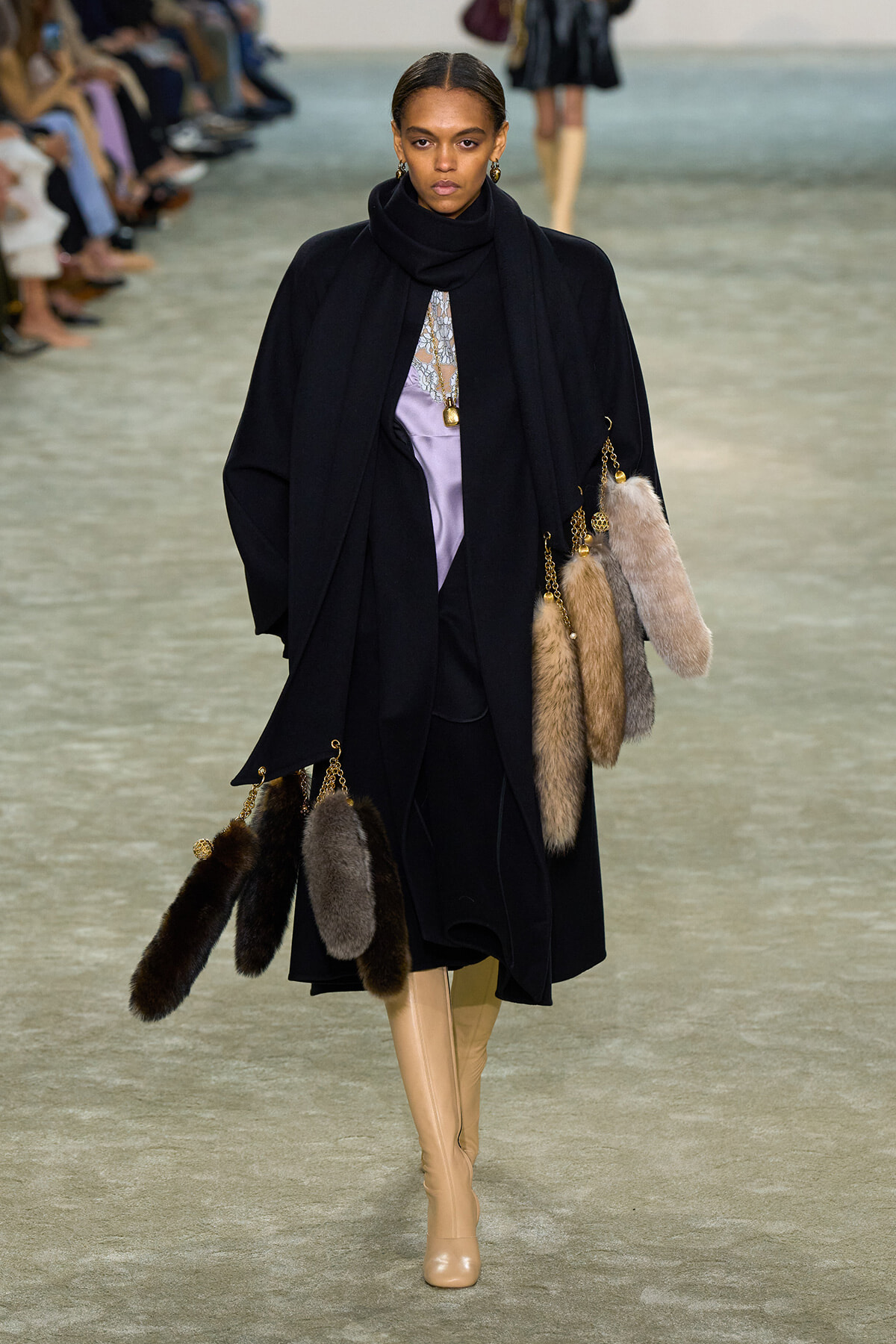 Model on runway in a long black coat with a scarf, lavender blouse, gold jewelry, and multiple fur tassel accessories; beige knee-high boots.