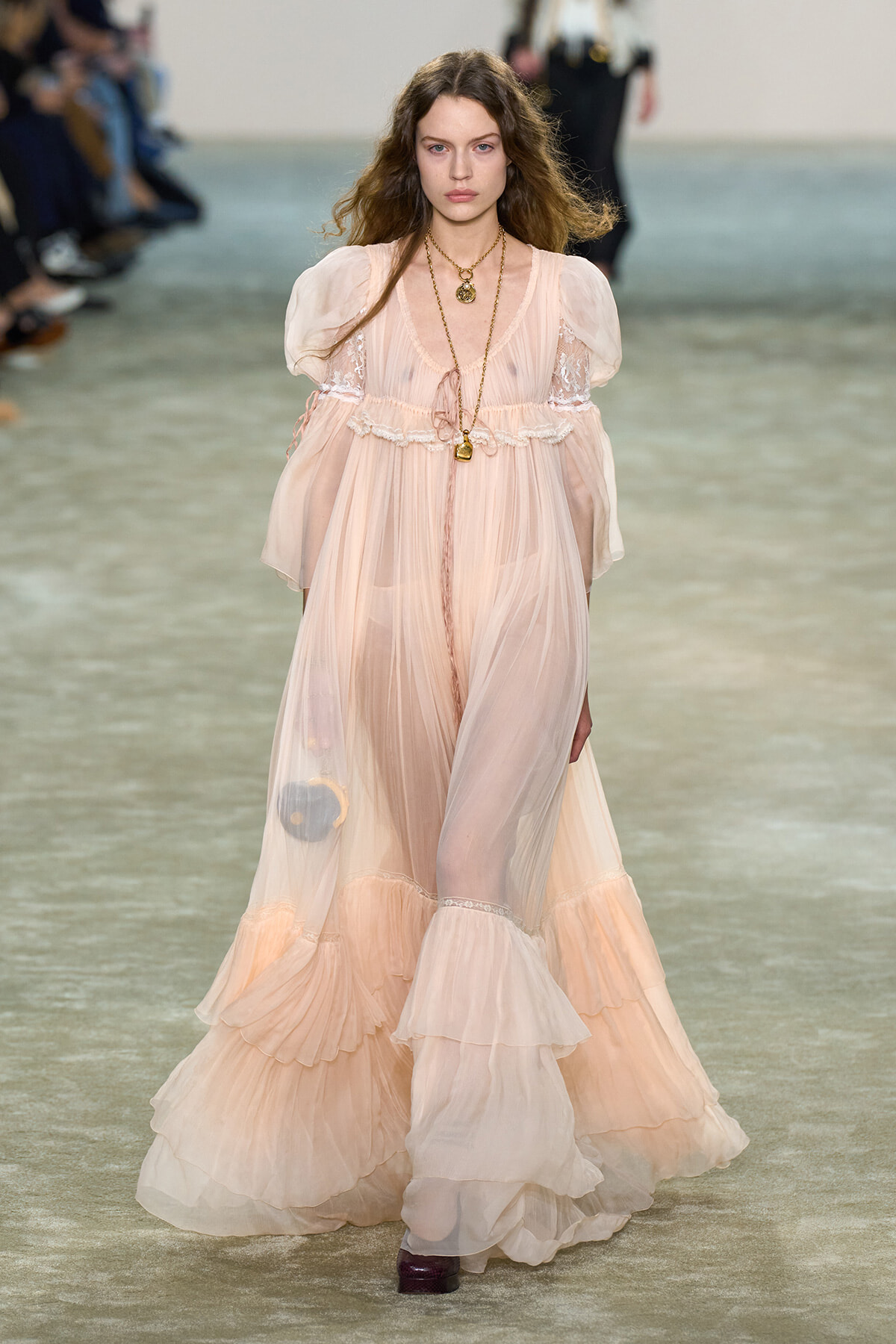 Model walks the runway in a long, pale pink sheer gown with puff sleeves, lace detailing, and layered ruffles, accessorized with gold necklaces.