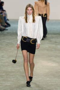 Model walking a runway in an ivory lace blouse with a high ruffled neck, a black mini skirt, and a wide gold-buckle belt, carrying a small round black handbag on a chain.