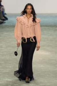 Model walks the runway in a peach ruffled blouse with a high neckline and voluminous sleeves, paired with a long black sheer skirt and a wide gold belt, holding a small black purse on a chain.