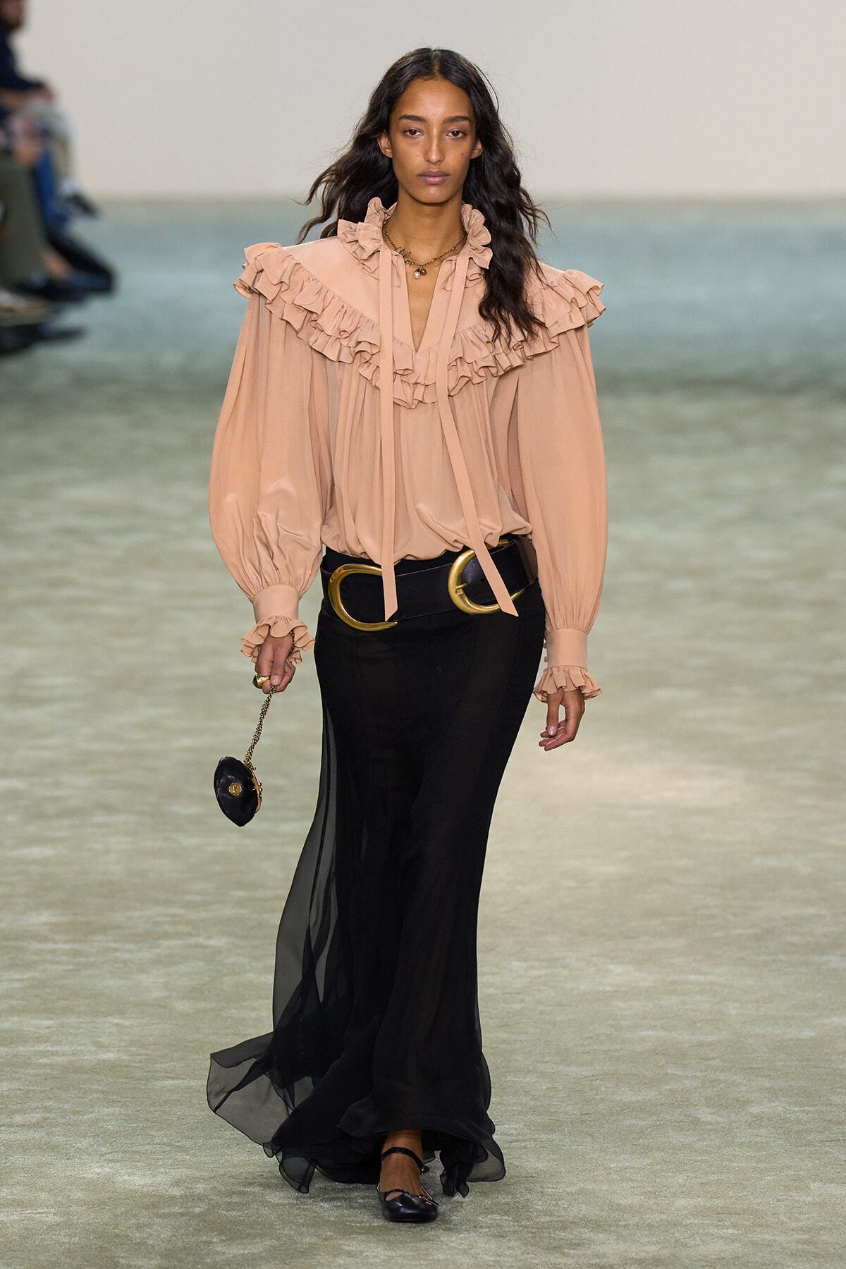 Model walks the runway in a peach ruffled blouse with a high neckline and voluminous sleeves, paired with a long black sheer skirt and a wide gold belt, holding a small black purse on a chain.