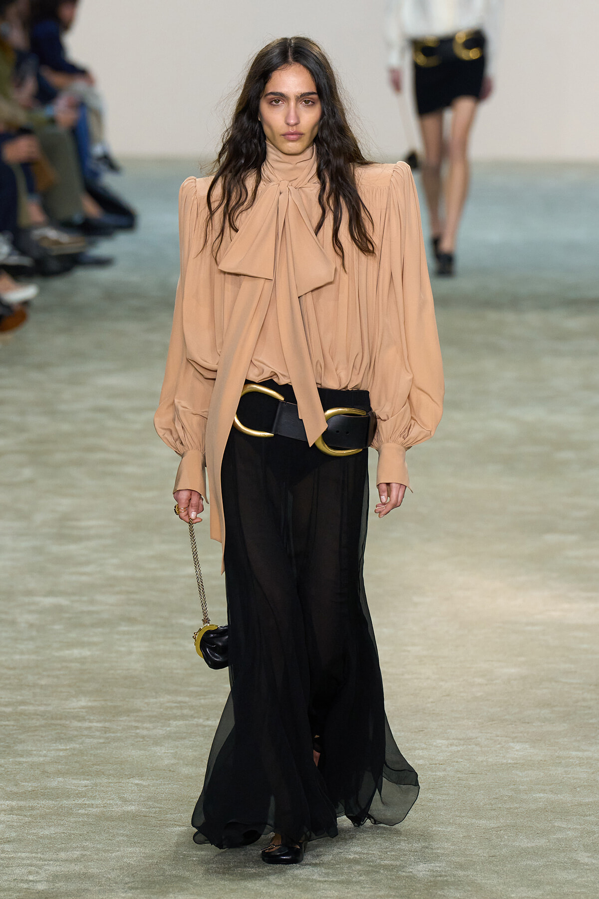 Female runway model wearing a peach blouse with a large bow, and sheer black wide-leg pants with a wide black belt and gold buckles.