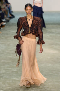 Model walking the runway in a bronze ruffled blouse with a deep V-neck and a peach flowing skirt, accessorized with gold jewelry and a burgundy bag forearm.