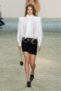 Model walking runway in a white billowy blouse with a pointed collar, paired with a black mini skirt and oversized gold-ring belt. A small chain purse dangles at her side.