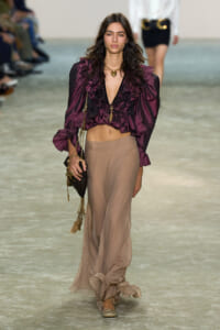 Runway model in a burgundy ruffled satin blouse paired with a beige sheer maxi skirt and gold jewelry.