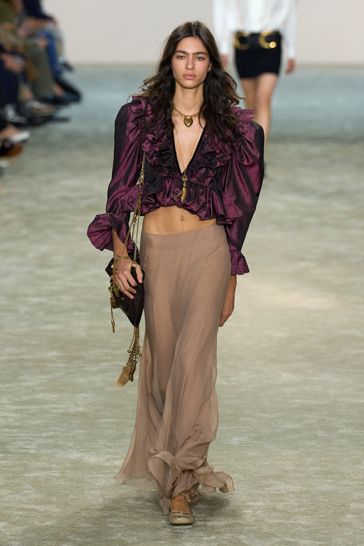 Runway model in a burgundy ruffled satin blouse paired with a beige sheer maxi skirt and gold jewelry.