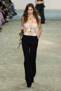 Model walks a fashion runway in a sheer white lace off-the-shoulder top, black wide-leg pants, and layered gold necklaces with a gold tassel bag.