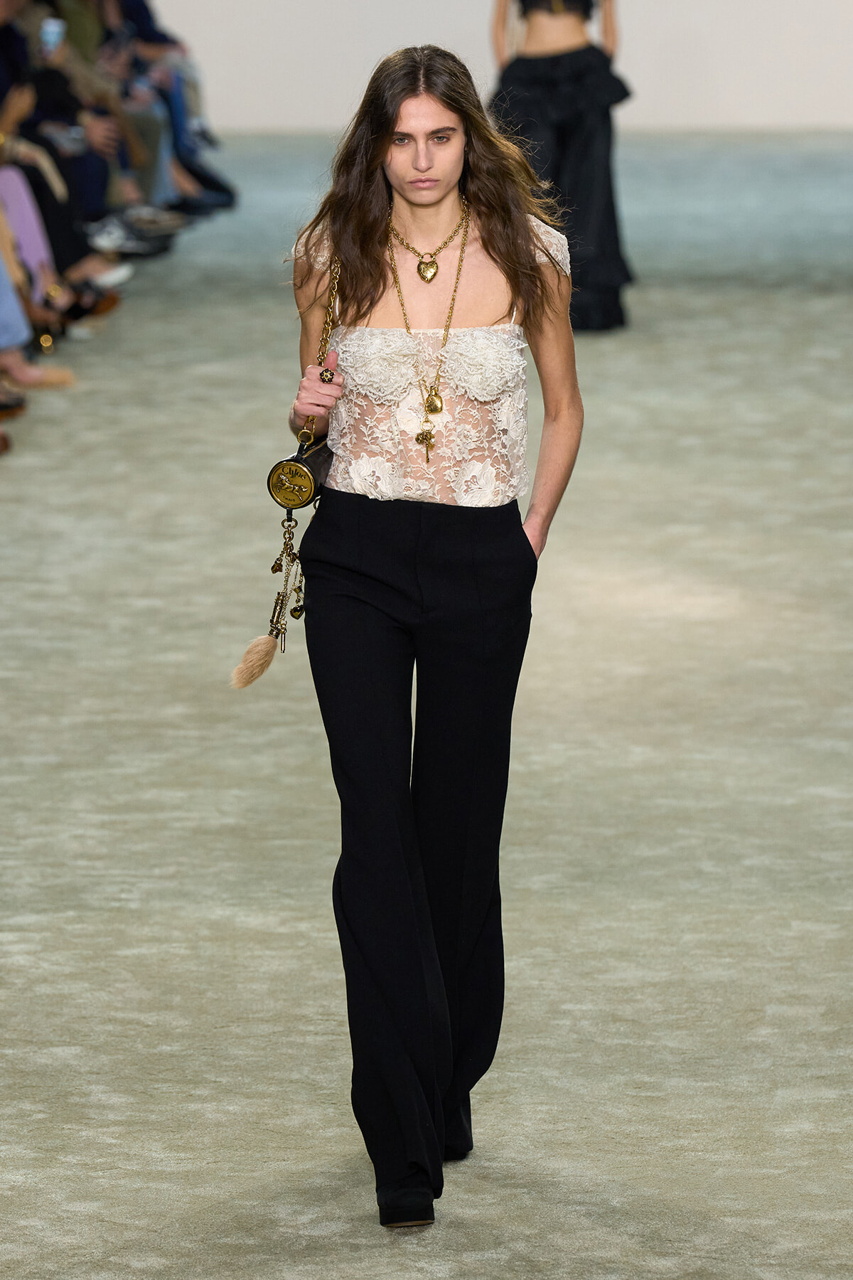 Model walks a fashion runway in a sheer white lace off-the-shoulder top, black wide-leg pants, and layered gold necklaces with a gold tassel bag.