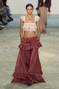 Runway model wearing a sheer white lace crop top and voluminous burgundy ruffled pants at a fashion show.