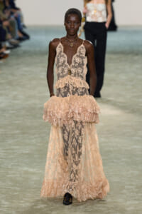 Black model walks the runway in a nude lace gown with a plunging neckline and tiered, ruffled skirt.