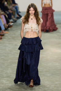 Model walking runway in a cream lace off-the-shoulder crop top and dark navy tiered maxi skirt with ruffles, brown sandals.