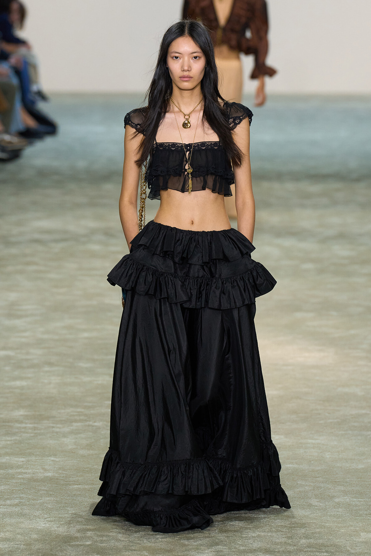 Female model on a runway wearing a black lace off-shoulder crop top with a layered, tiered maxi skirt and gold jewelry.