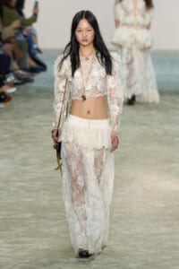 Model walking a runway in a white lace two-piece outfit with a cropped top and long sheer skirt, accessorized with gold jewelry and a small bag.