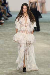 Model walks the runway in an ivory sheer lace gown with layered ruffles and a long gold pendant necklace.