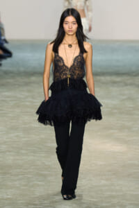 Model on runway wearing a black lace sheer top with tiered peplum over black pants, layered gold necklaces.