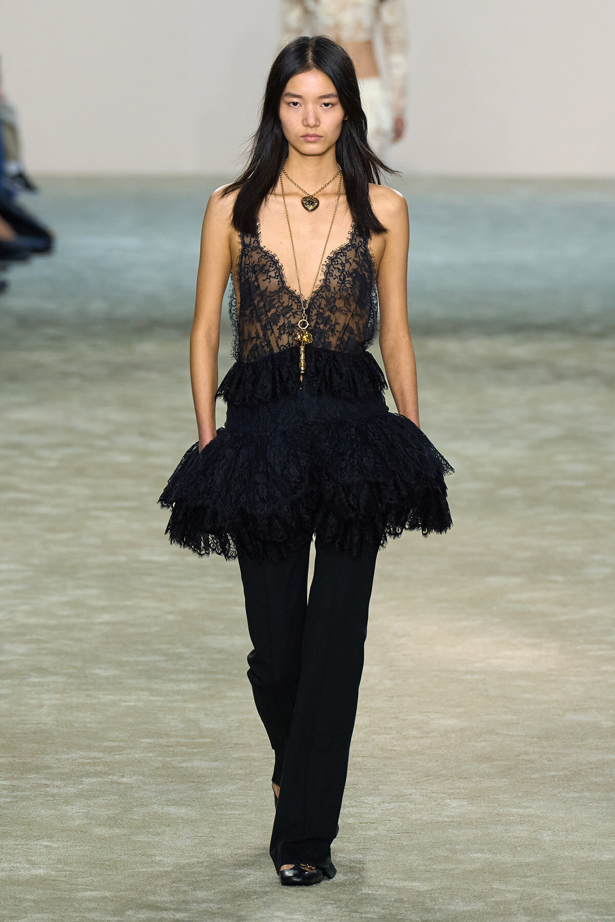 Model on runway wearing a black lace sheer top with tiered peplum over black pants, layered gold necklaces.
