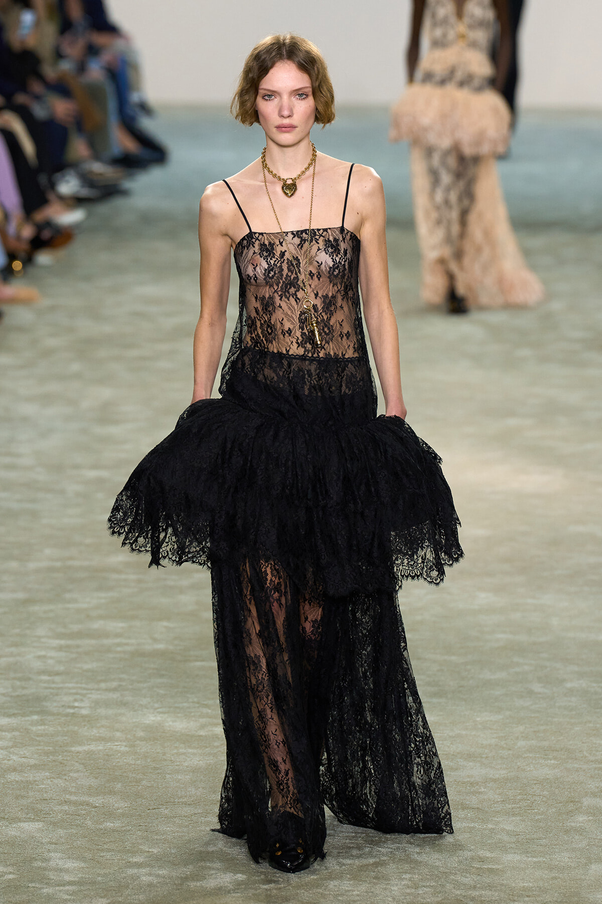 Model walking a runway in a black lace sheer-bodice gown with a layered lace skirt and gold necklace