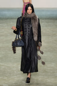 Model walks the runway in a long black leather coat with a thick gray fur scarf, carrying a navy handbag with a gold chain,