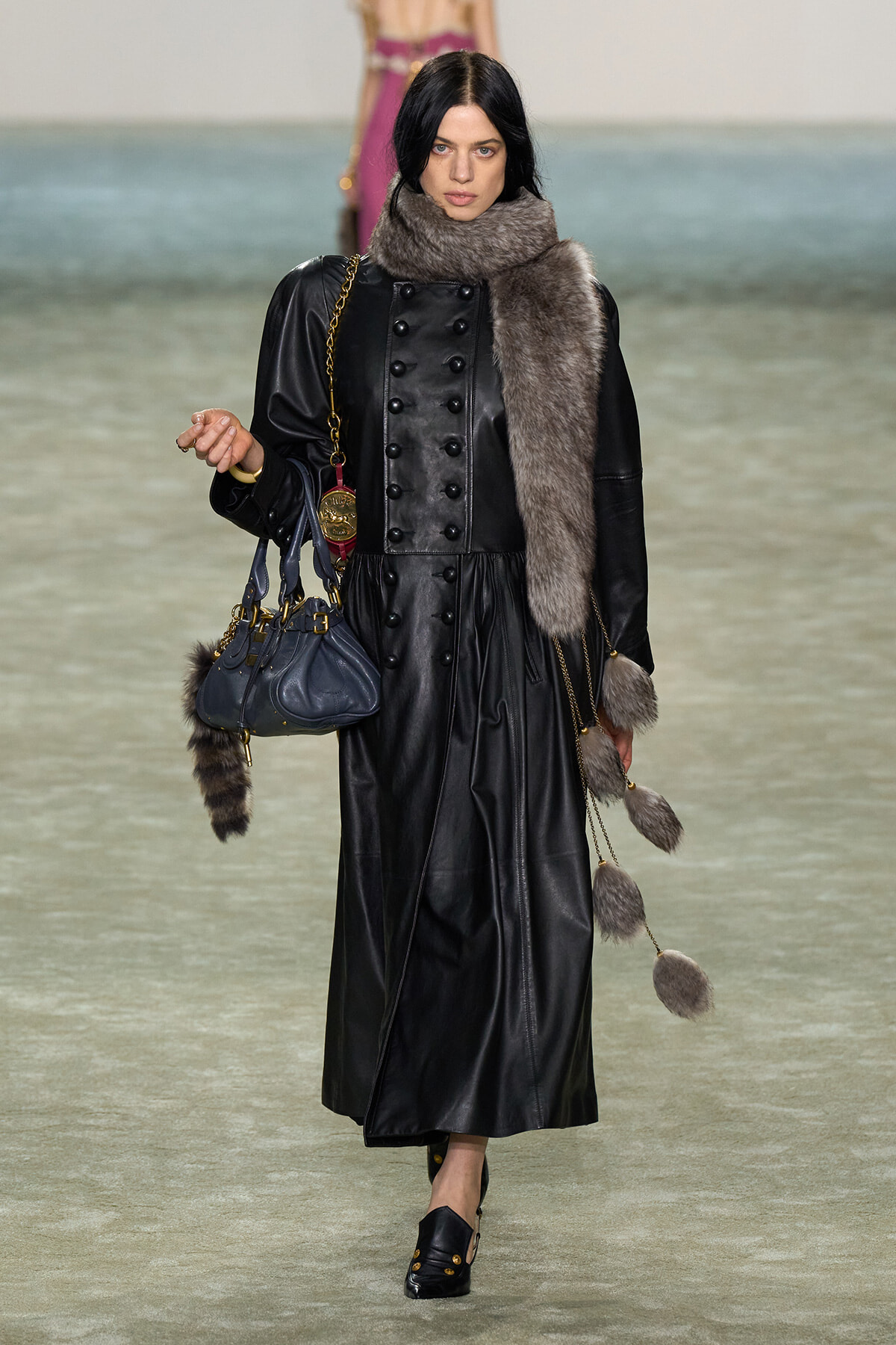 Model walks the runway in a long black leather coat with a thick gray fur scarf, carrying a navy handbag with a gold chain,