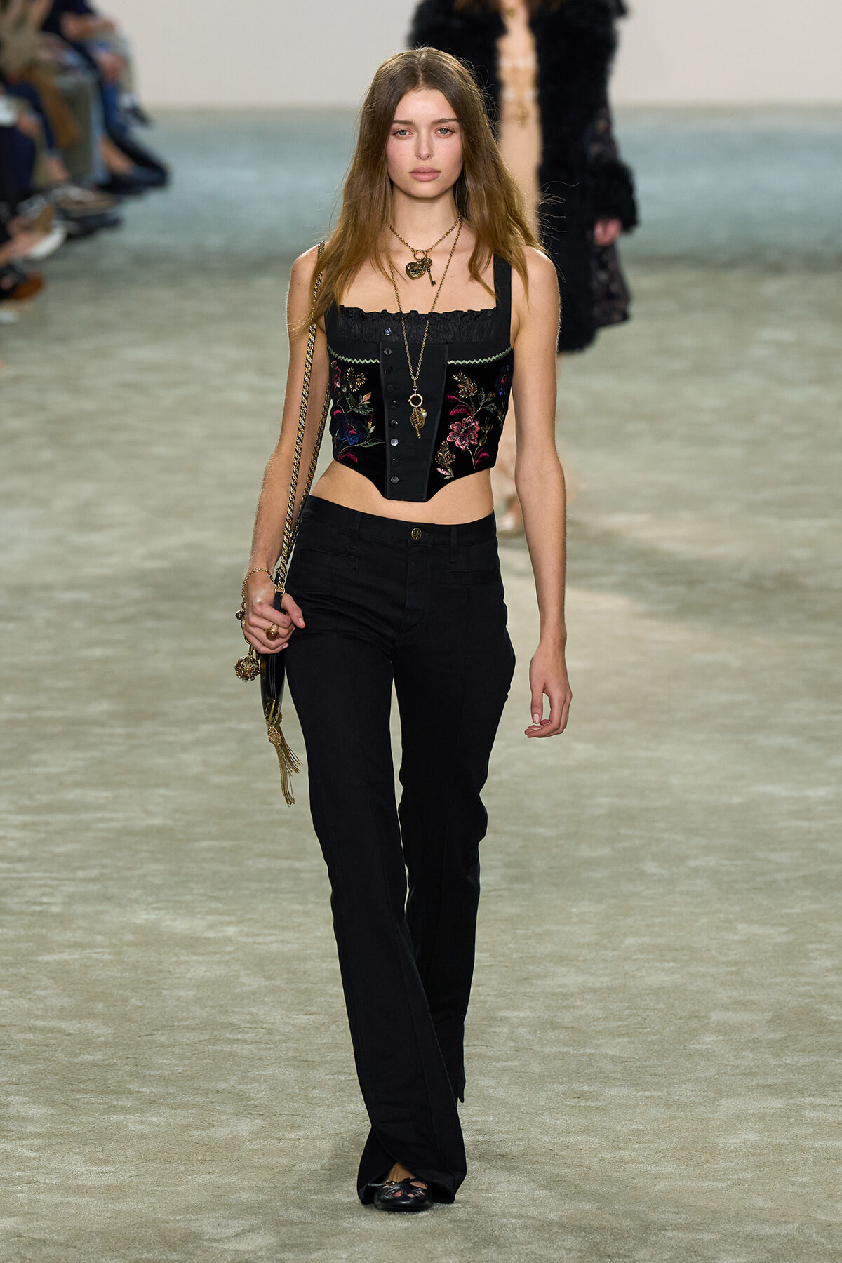 Model on a runway wearing a black embroidered cropped corset top and black pants, with layered gold necklaces and a chain-strap bag down her side.