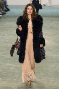 Model walking the runway in a long beige lace-trim dress under a black faux-fur coat, accessorized with gold jewelry and a burgundy handbag with a fur keychain accessory