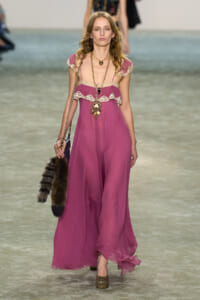 Model walking a fashion runway in a magenta, off-shoulder chiffon dress with cream lace trim and layered necklaces, carrying a fur-tailed bag.