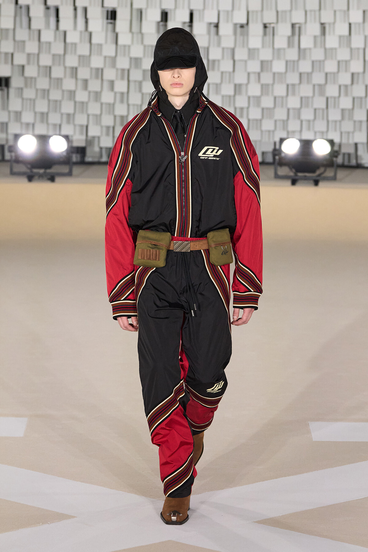 Model on a runway wearing a black and red hooded jumpsuit with striped piping and a brown utility belt.