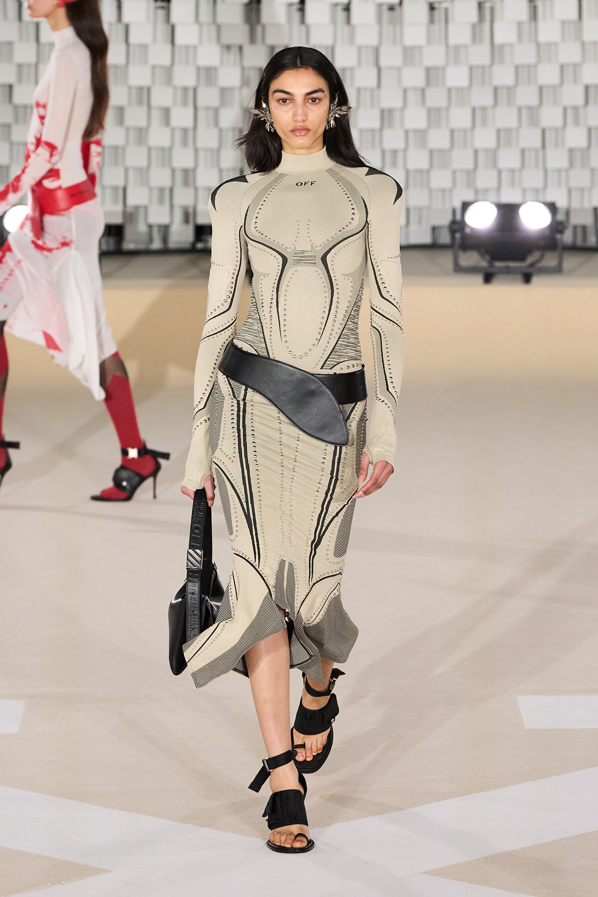 Model walks a fashion runway wearing a beige sculpted-knit dress with black stitched patterns and a wide black belt bag, carrying a black shoulder bag.