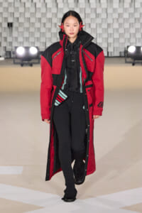 Model walks the runway in a red and black color-blocked coat with a high collar and zippers.