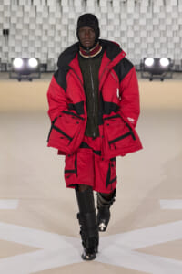 Model walking a runway in a red and black insulated parka with multiple pockets, matching red shorts, and tall black boots.