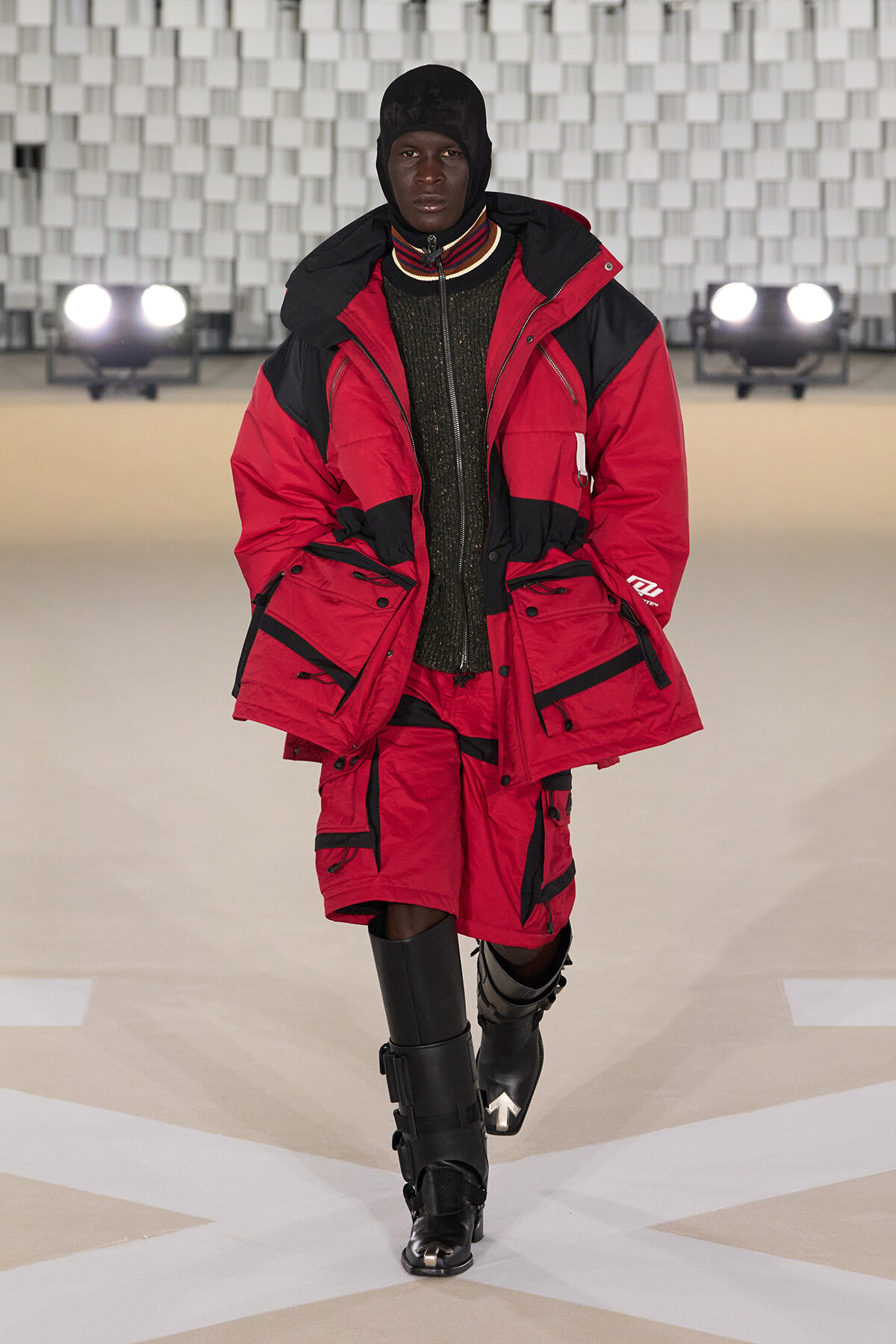 Model walking a runway in a red and black insulated parka with multiple pockets, matching red shorts, and tall black boots.
