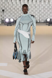 Model walks a fashion runway in a pale blue sculpted dress with riveted detailing and a large white belt, carrying a blue bag and wearing black strappy sandals.