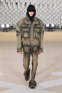 Model walking the runway in a distressed olive-green studded military-style jacket and pants, with a black hood and belt.