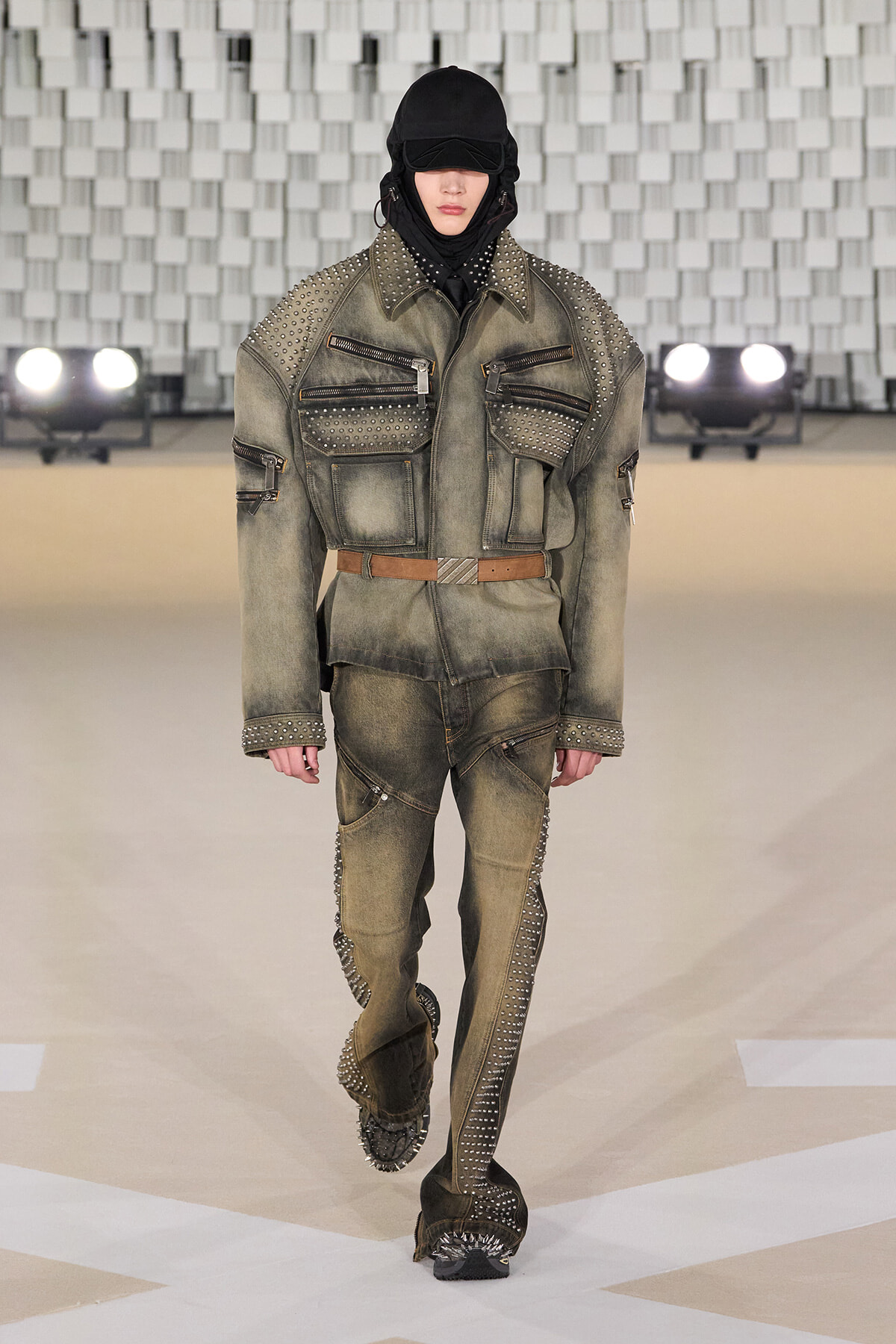Model walking the runway in a distressed olive-green studded military-style jacket and pants, with a black hood and belt.